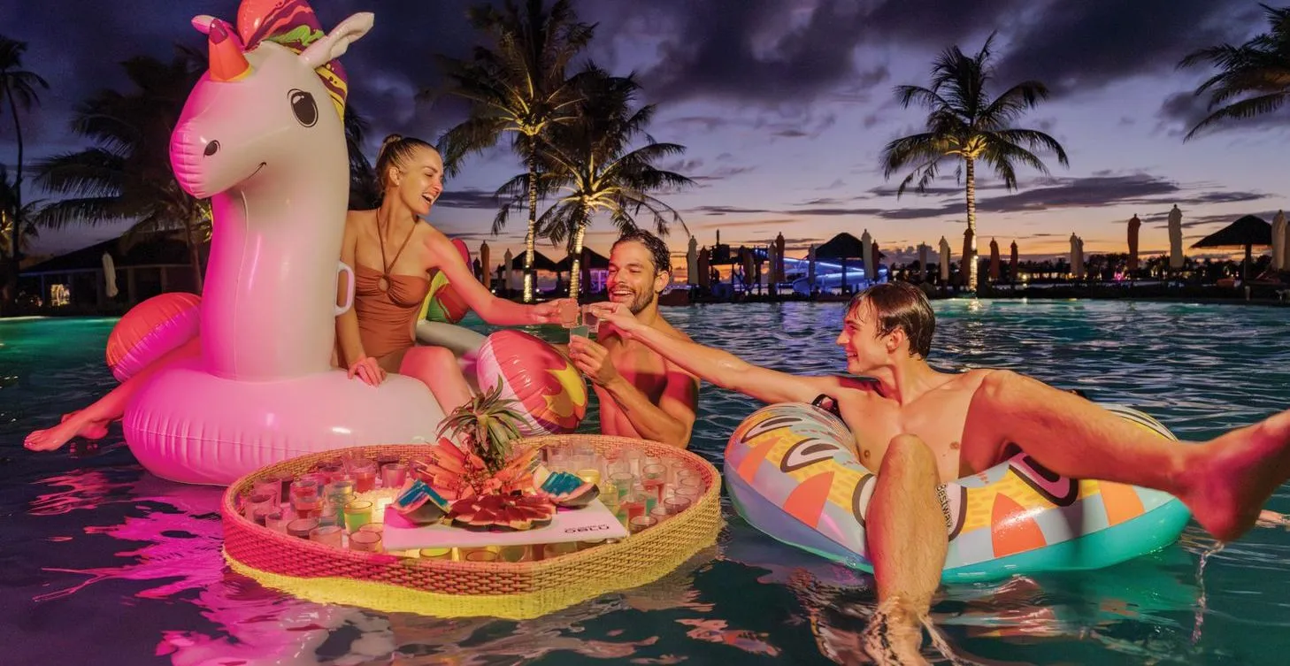 3 friends on an inflatable unicorn and ring in the pool at sunset, a tray of fruit floating between them.