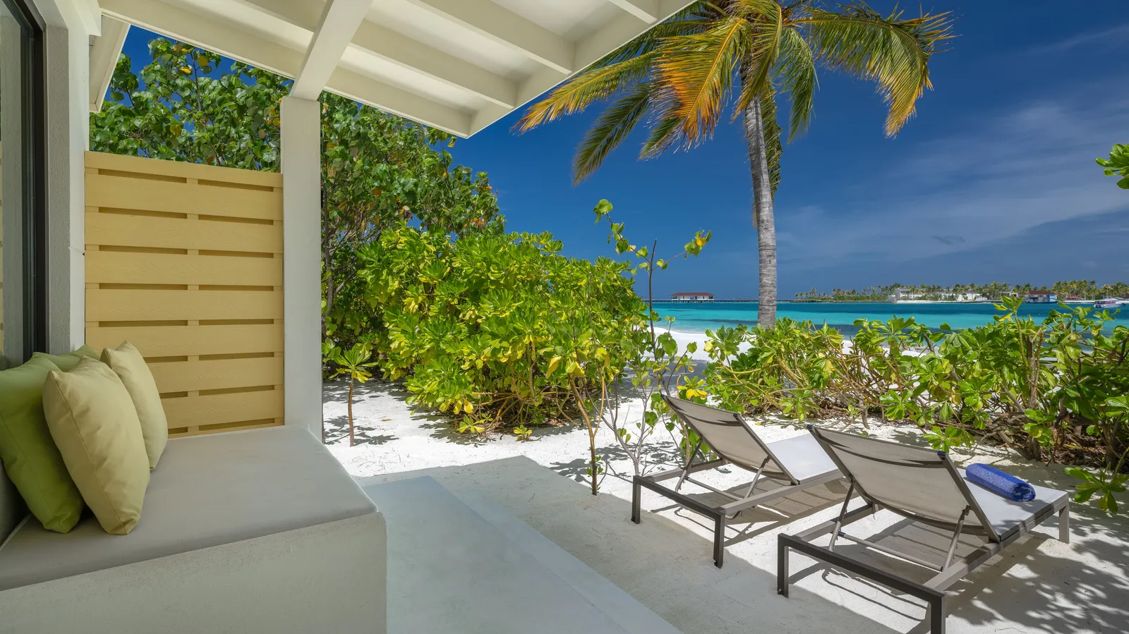 The veranda of a Beach Villa: the daybed, 2 loungers on the sand and, beyond the shrubs, the lagoon.