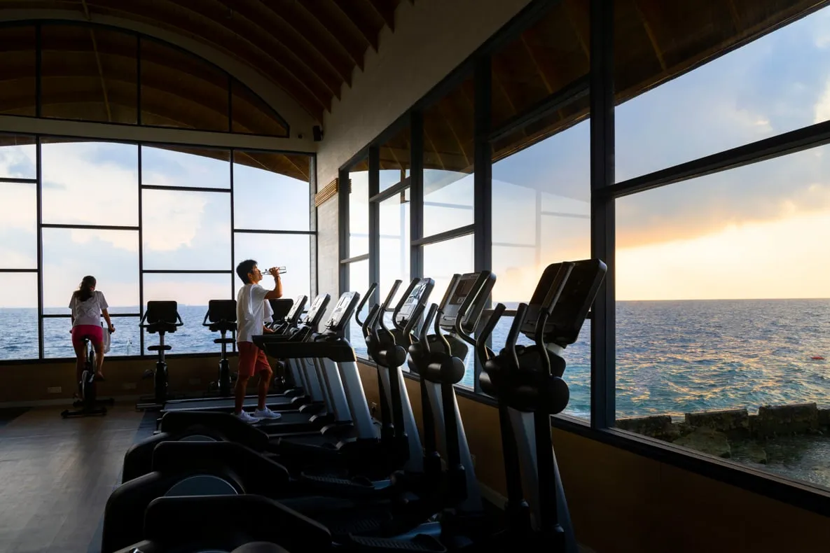 The gym with its windows on the sea at sunset, a row of treadmills and 2 people training.