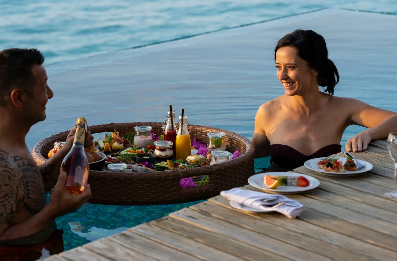 A couple in their villa pool with a floating breakfast in a large basket, among fruit, juices and a bottle of sparkling wine.
