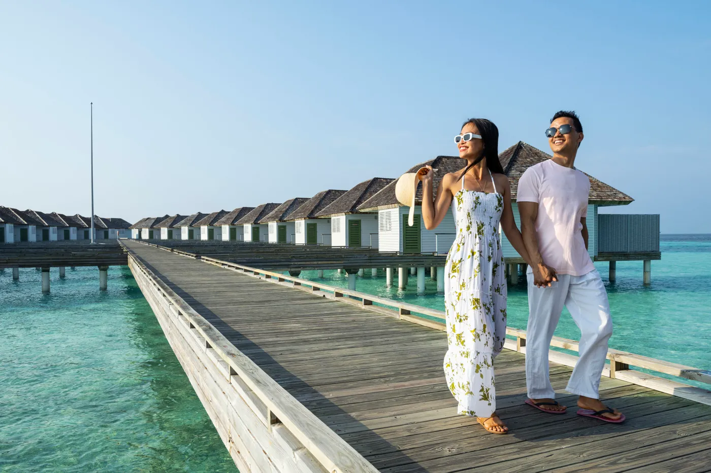 A couple walk hand in hand along the wooden jetty, past the row of water villas with their shingled roofs.