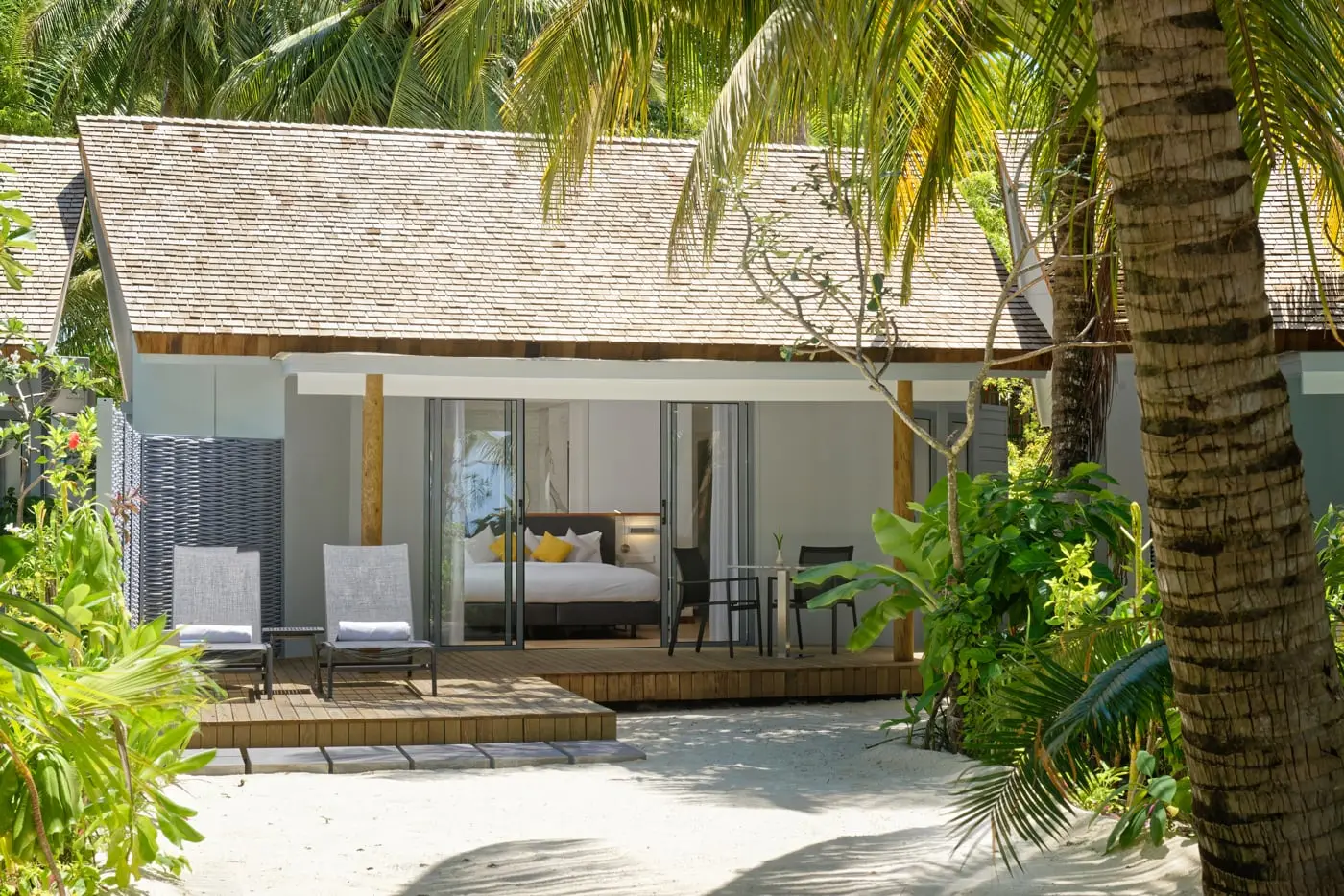 A Beach Villa among the palms, with its wooden deck, 2 loungers and the glass doors open to the bed.