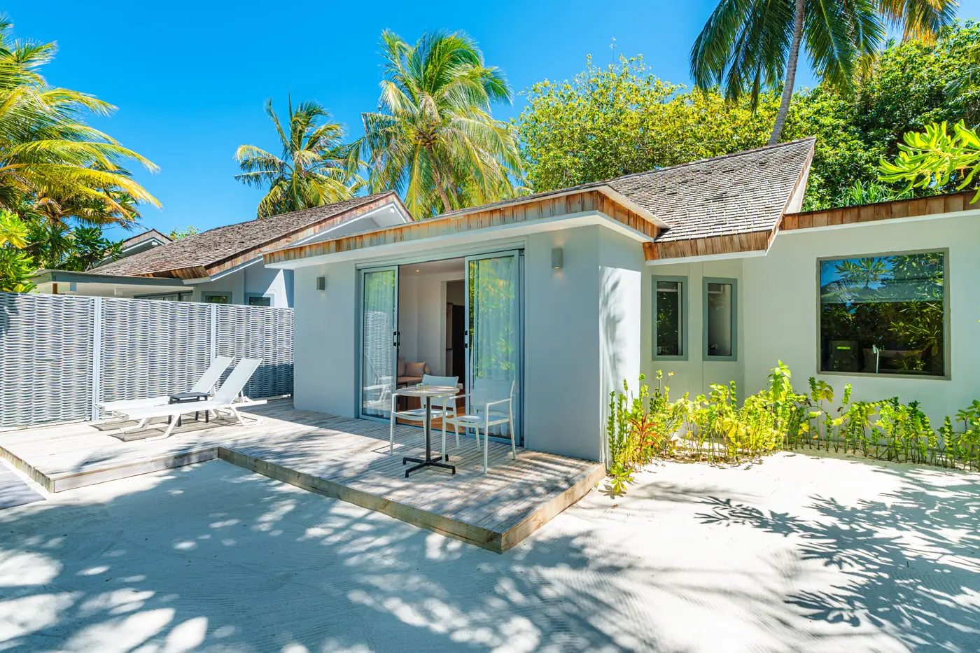 The white Family Beach Villa among the palms, its deck on the sand with loungers, a table and chairs.