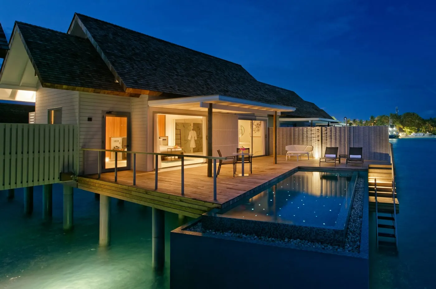 A Sunset Over Water Villa with Private Pool in the evening, lit inside, with its pool glowing above the dark water.