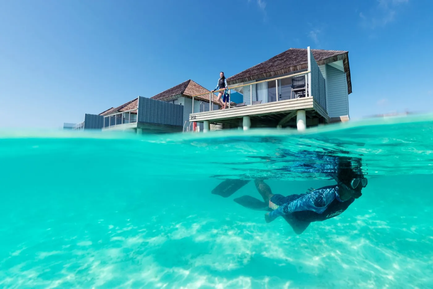 Half above and half below the surface: a swimmer in mask and fins in the lagoon in front of a water villa.