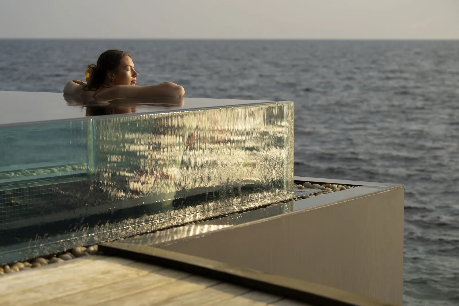 A woman resting on the glass edge of a spa hot tub that hangs over the sea at dusk.