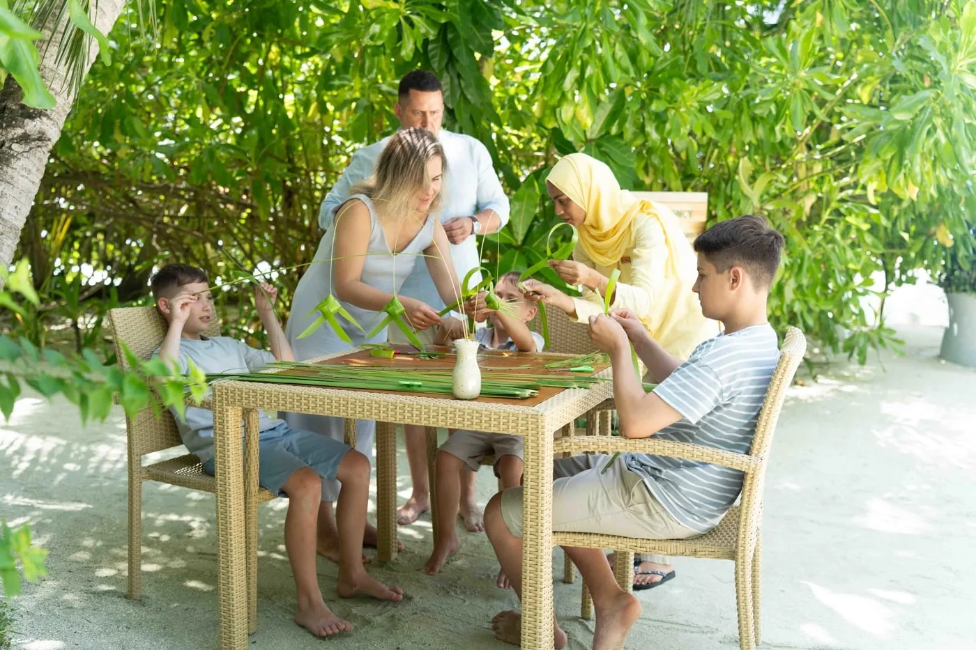 A family with 2 boys weave palm fronds at an outdoor table in the shade of the trees, on the sand.