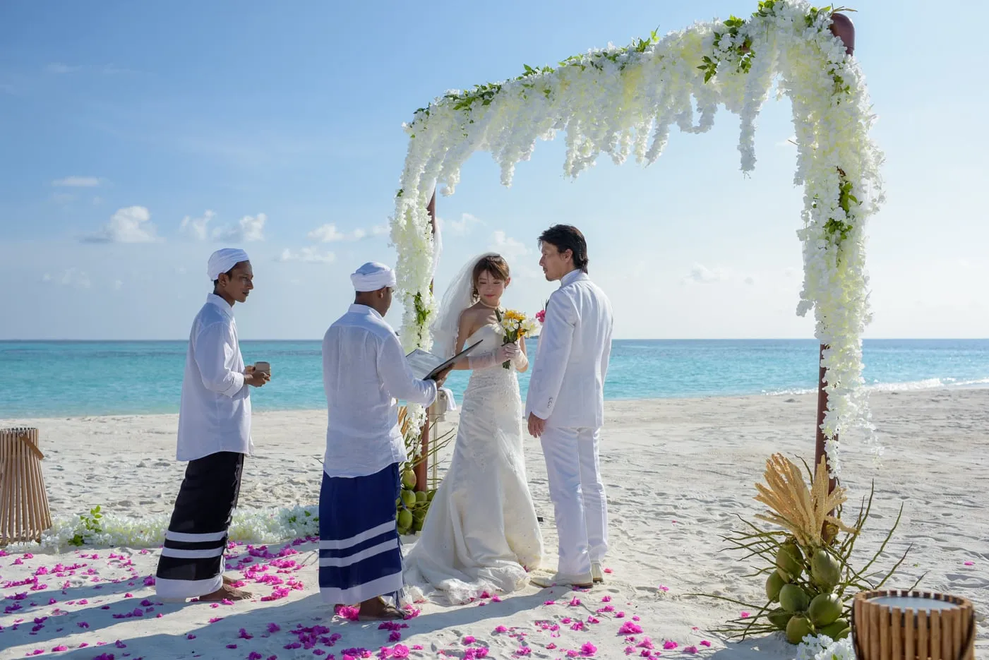 A couple in white before the arch of flowers on the beach, with 2 men in sarongs for the vow renewal ceremony.