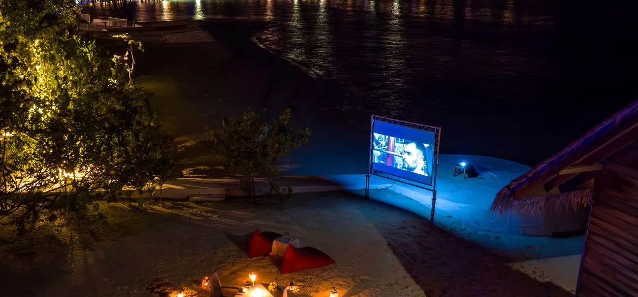 The beach cinema at night: the lit screen, the cushions and the candles on the sand.