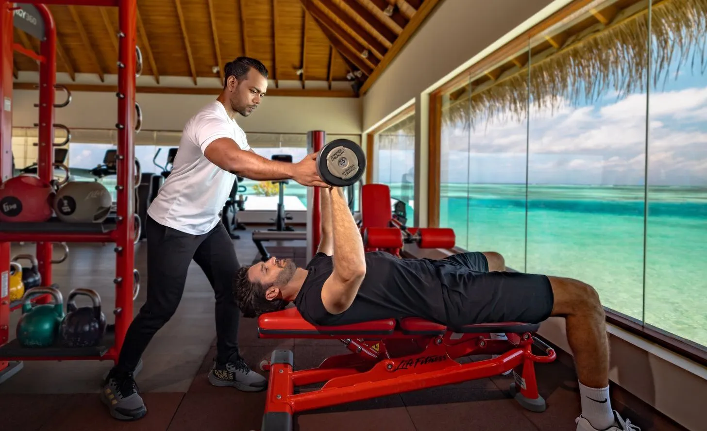 The overwater gym: a trainer spotting a guest on the bench, with the lagoon beyond the glass.