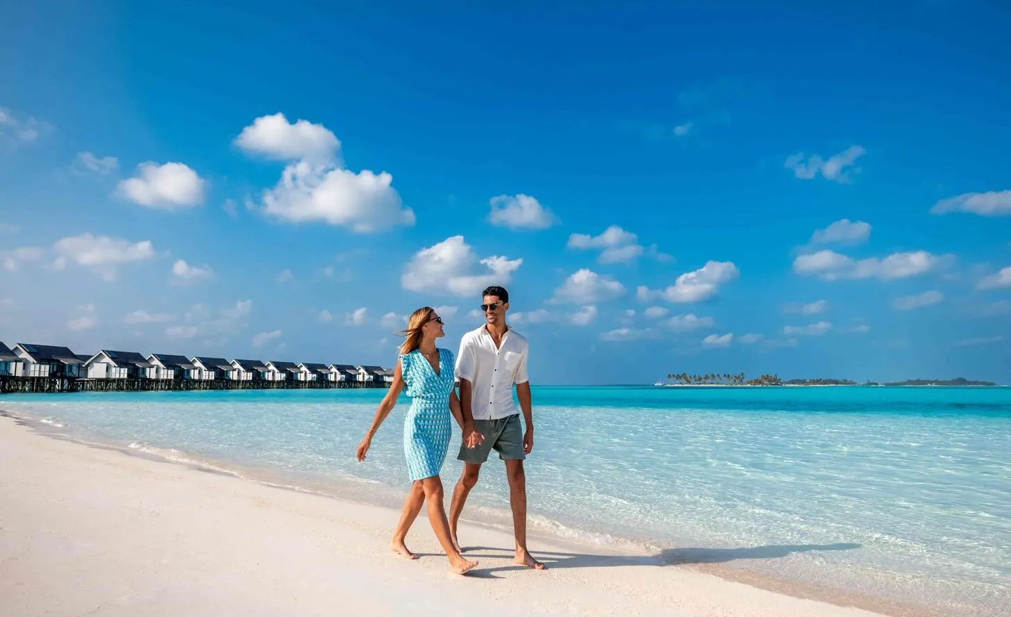 A couple walking on the white beach, with the water villas in a row behind them.