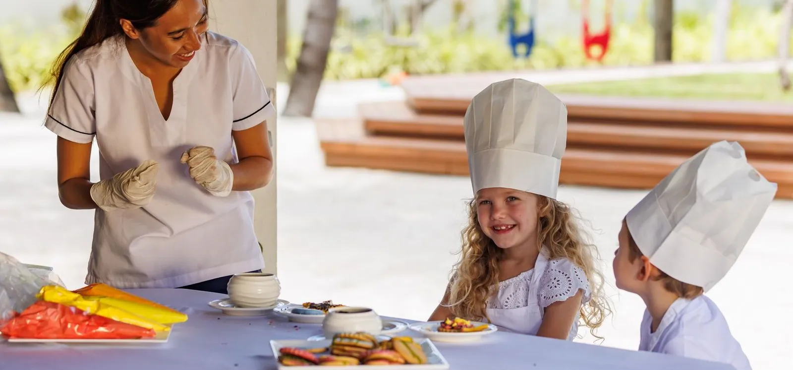 2 children in chef’s hats laughing at a table in the Kuda Koli Kids Club, while a carer prepares fruit.