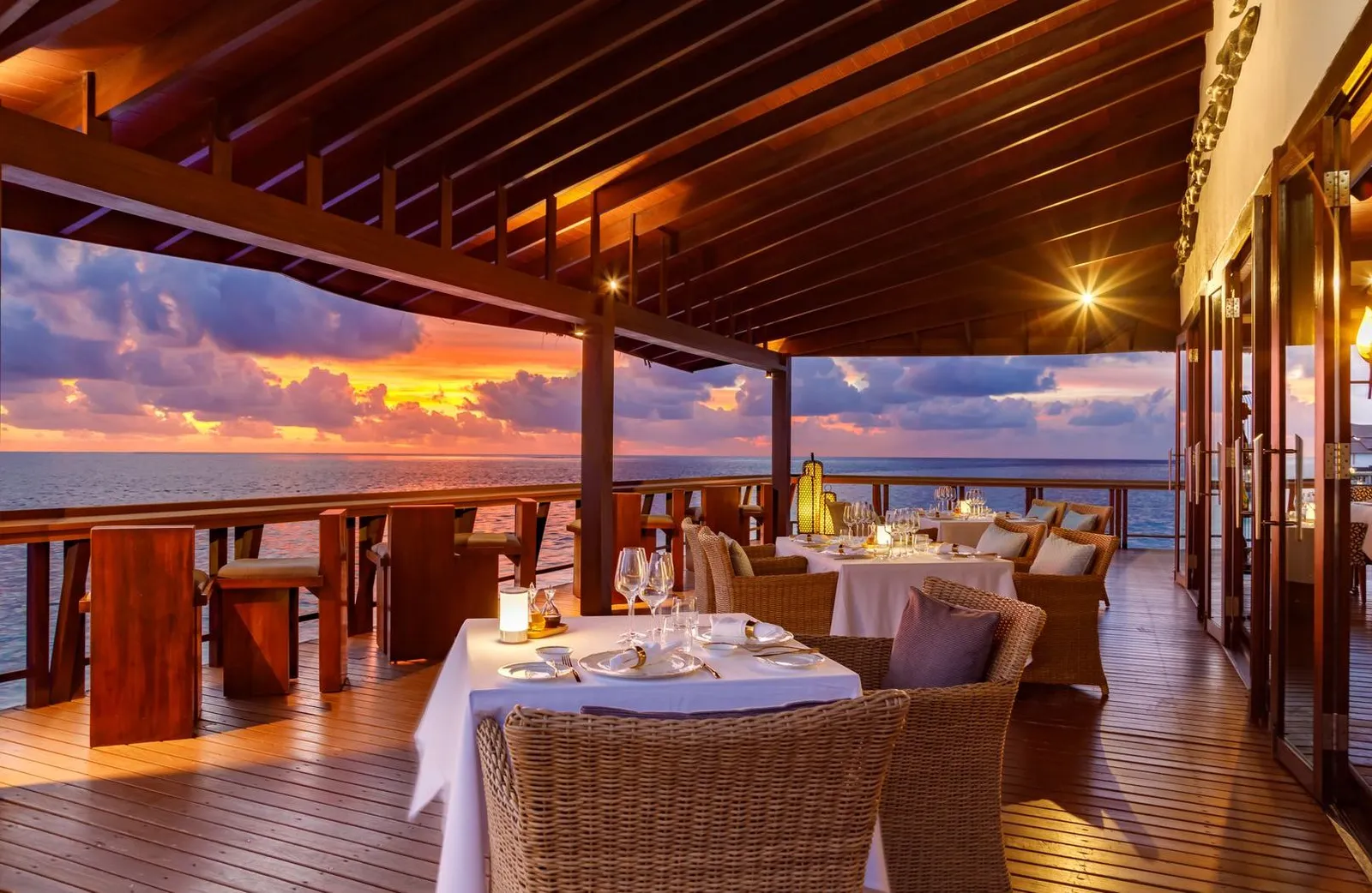ORIGINƎ’s terrace at sunset, tables laid under the timber roof and the sea beyond the railing.