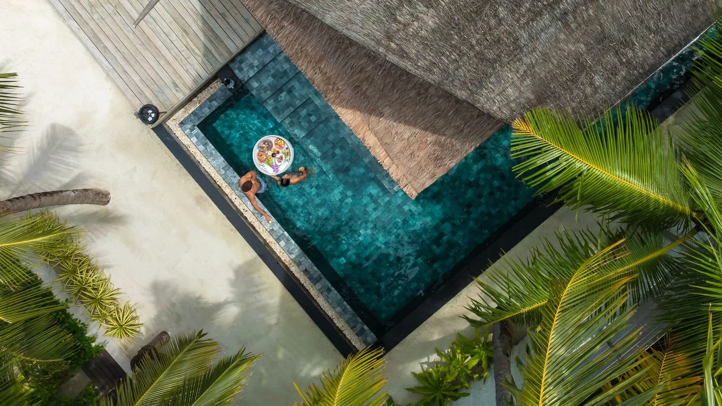 An Earth Pool Villa Sunset’s L-shaped pool from above, a guest floating among the palms.