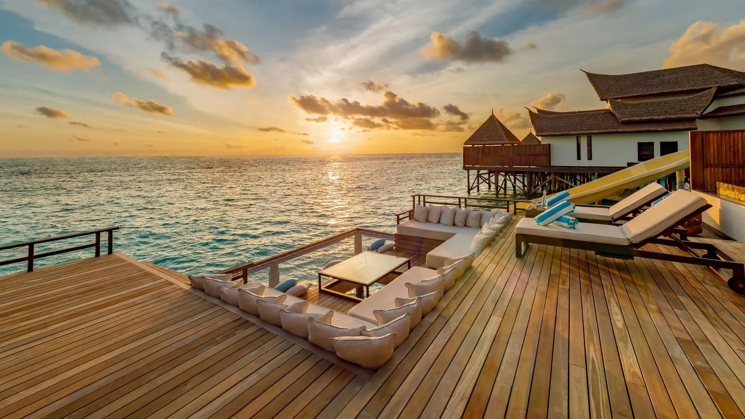 A water villa’s timber deck at sunset, with its outdoor sofa and loungers facing the sea.