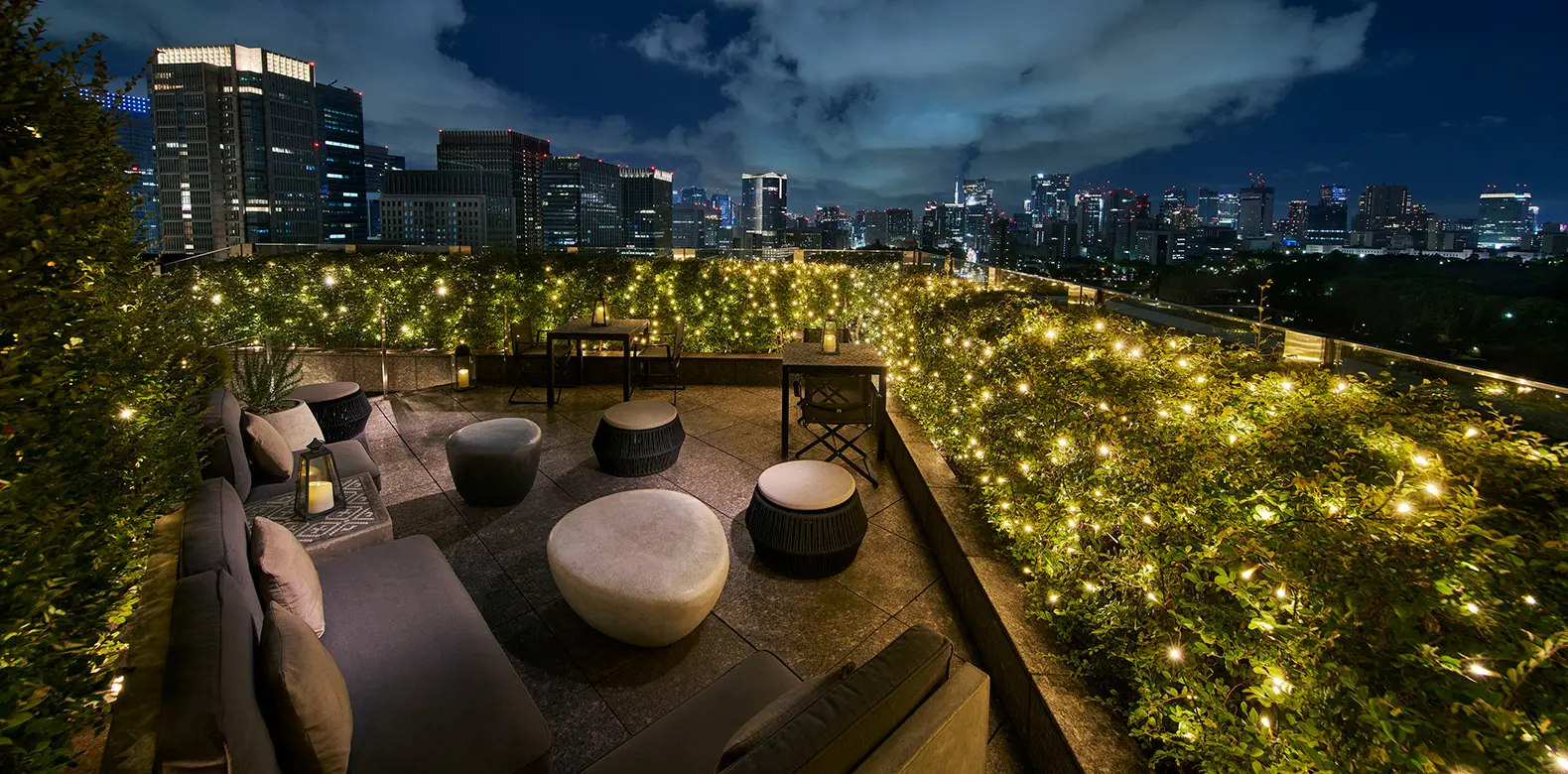 The Club Lounge terrace at night, among plants and fairy lights, with the lit towers around it.