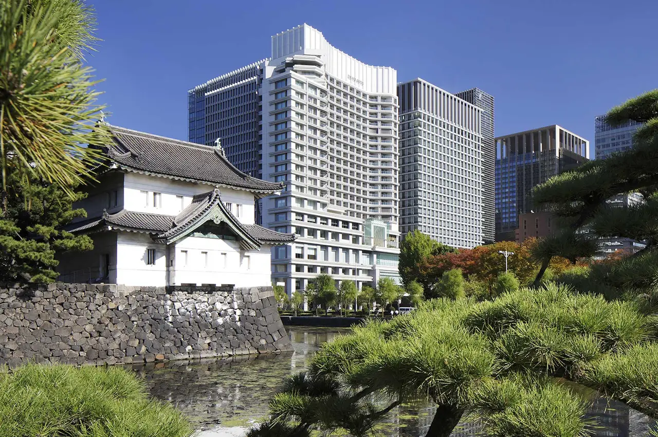 The white Tatsumi watchtower on its stone walls among the pines of the palace gardens, with the hotel’s building behind it.