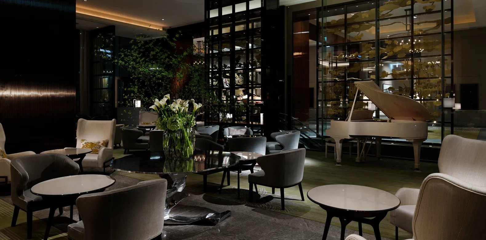 The Palace Lounge in the evening, with armchairs, low tables and the grand piano by the windows.