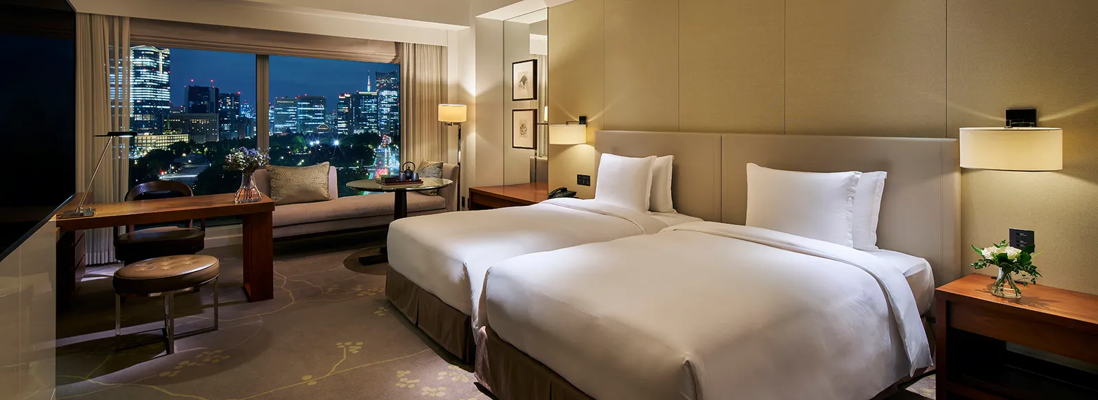 A Grand Deluxe twin room in the evening, the desk under the window and the city lit up.