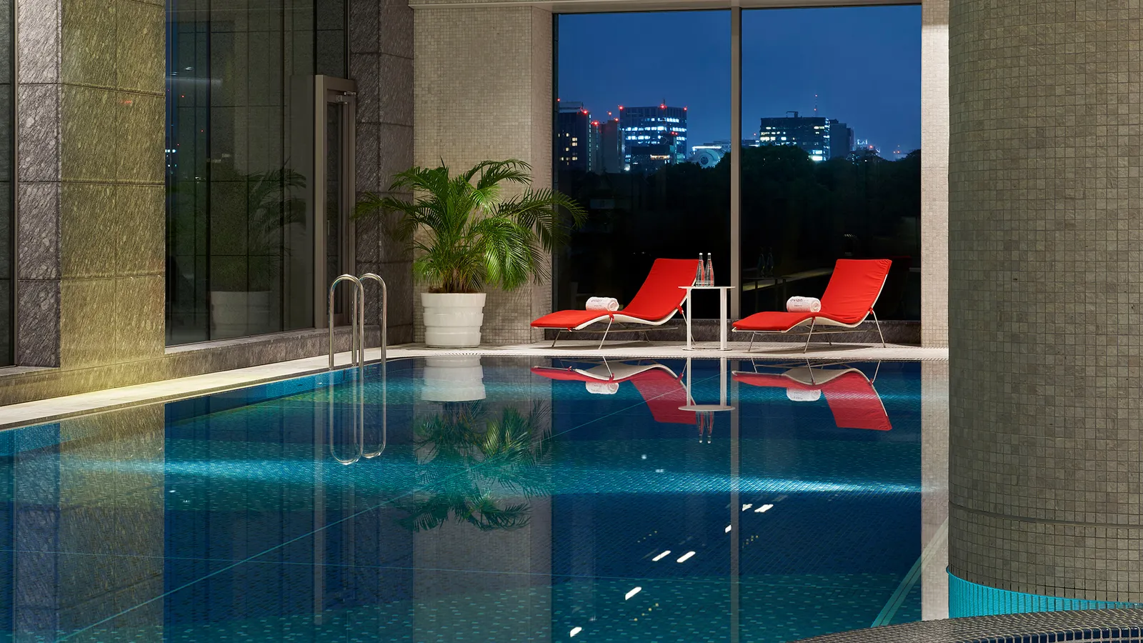 The indoor pool at dusk, with red loungers at its edge and the window onto the lit towers of Marunouchi.
