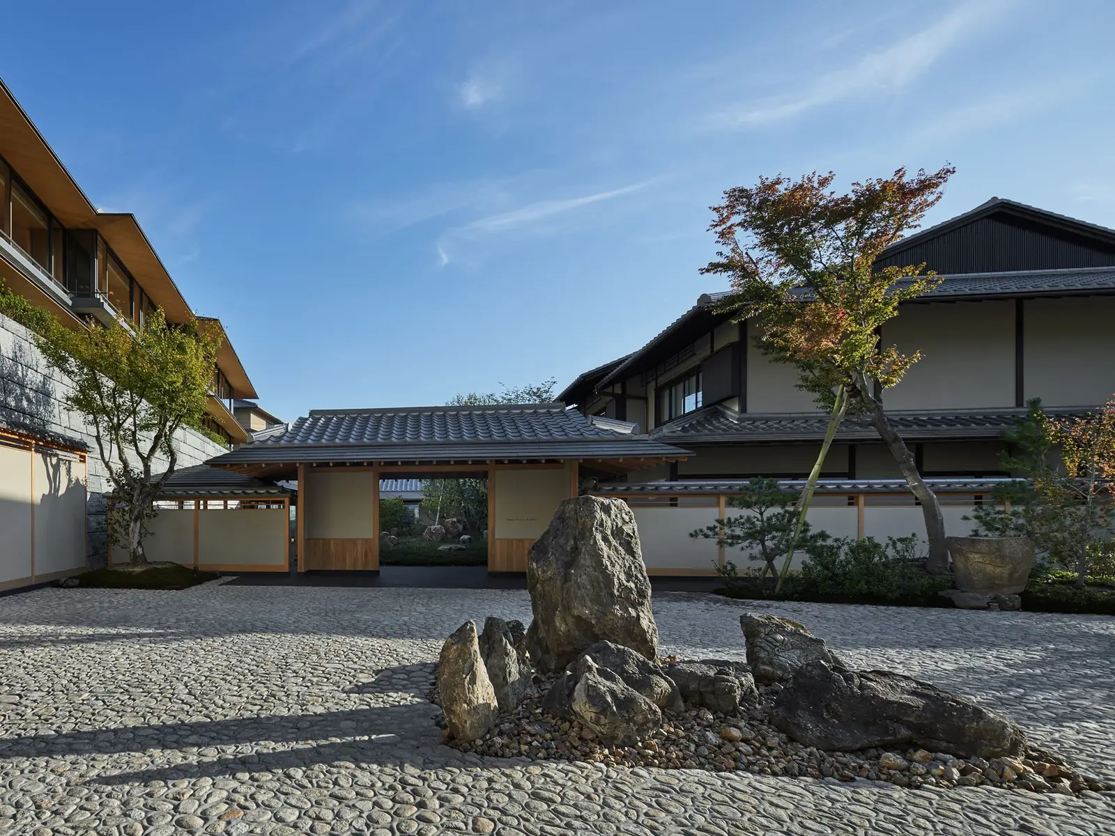 The garden of raked gravel and rocks before the roofed gate, with a maple and the hotel’s pavilions.