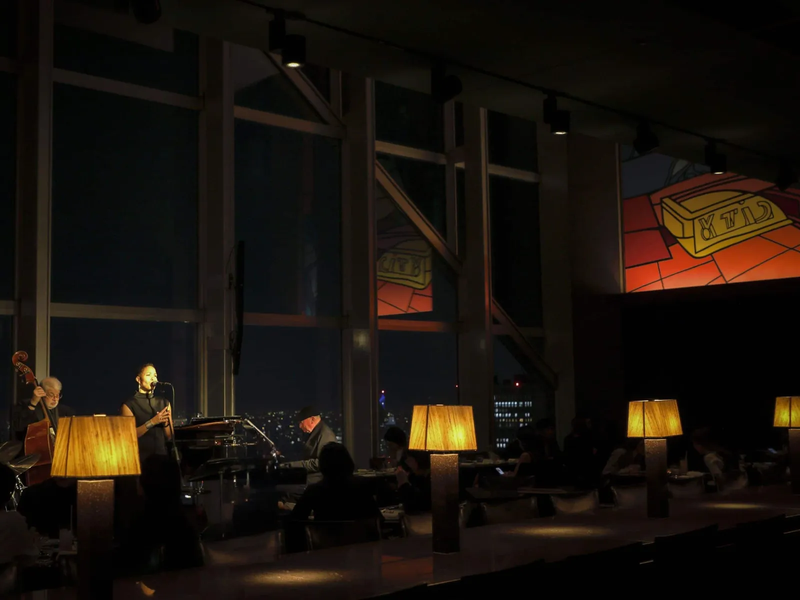 New York Bar at night: a singer, a double bass player and a pianist perform in front of the windows, among guests at their tables and the lit lamps.