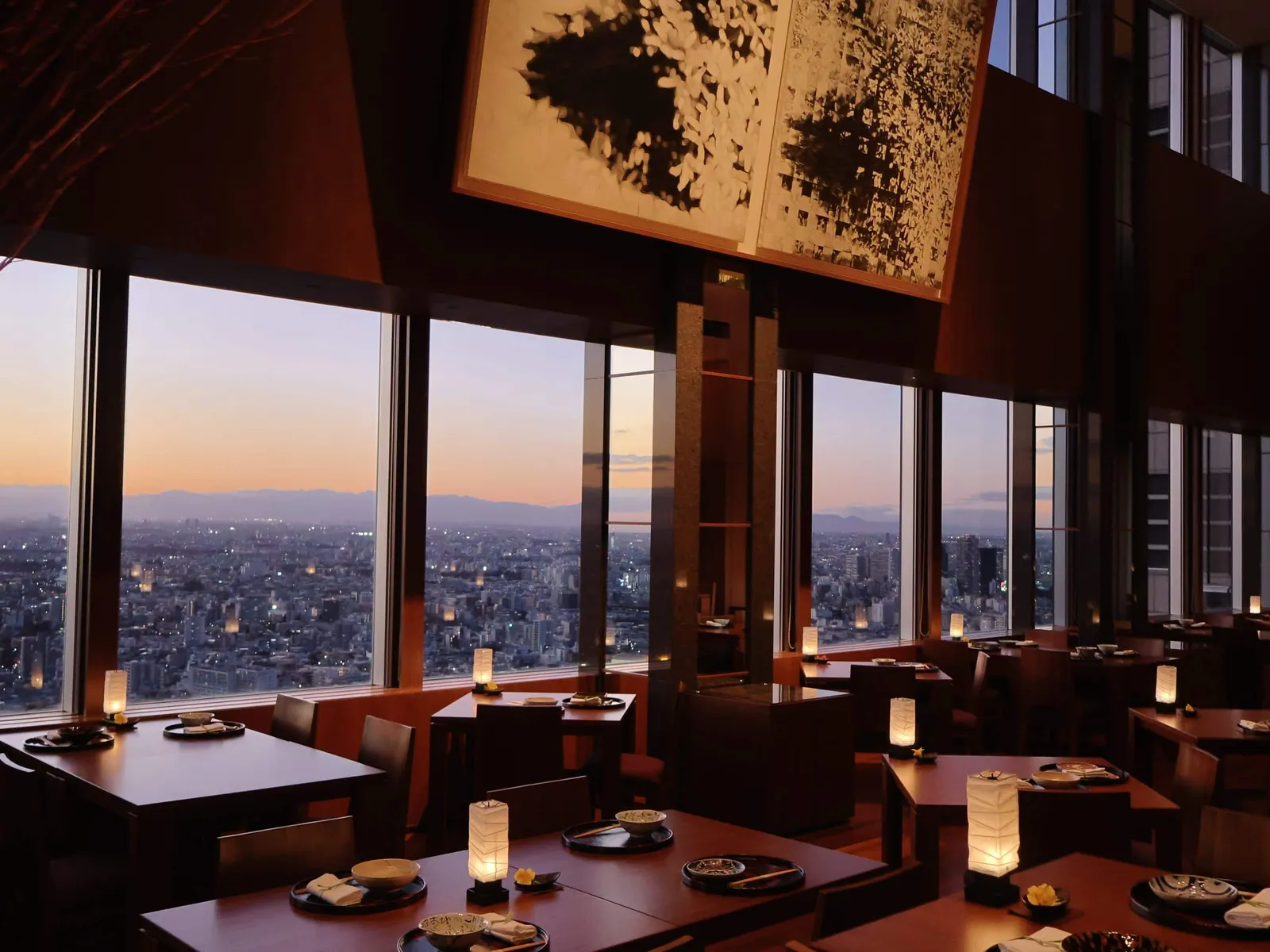 Kozue at sunset: wooden tables with paper lamps lit, the windows over the city and a large black and white abstract picture.
