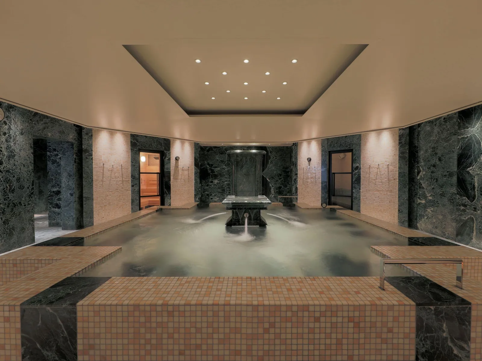 The spa’s whirlpool between dark green marble walls and sand-coloured mosaics, with a stone fountain in the middle.