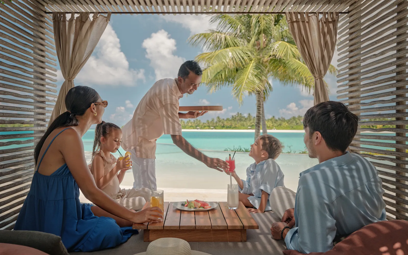 A family in a beach cabana, a waiter bringing drinks and the lagoon beyond the curtains