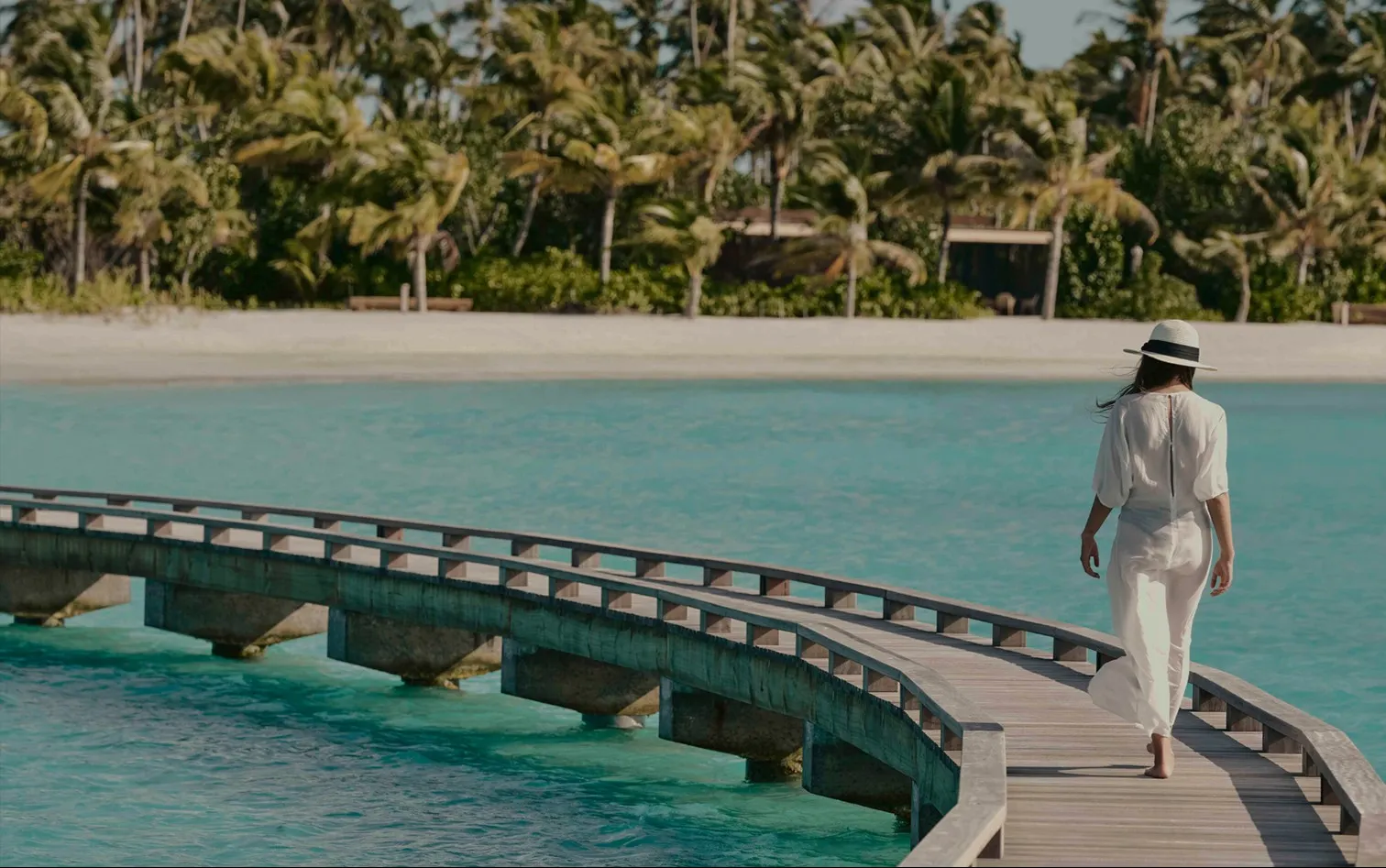 A woman in white walking the curved boardwalk over the lagoon, palms along the shore