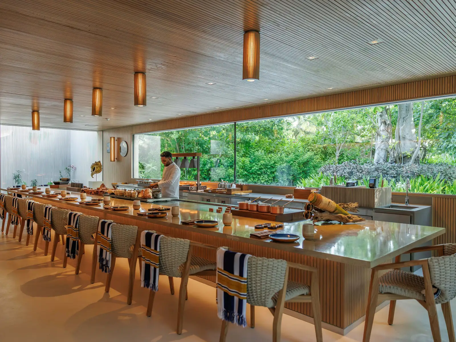 A restaurant’s long counter, chairs lined up before the open kitchen and greenery beyond the glass