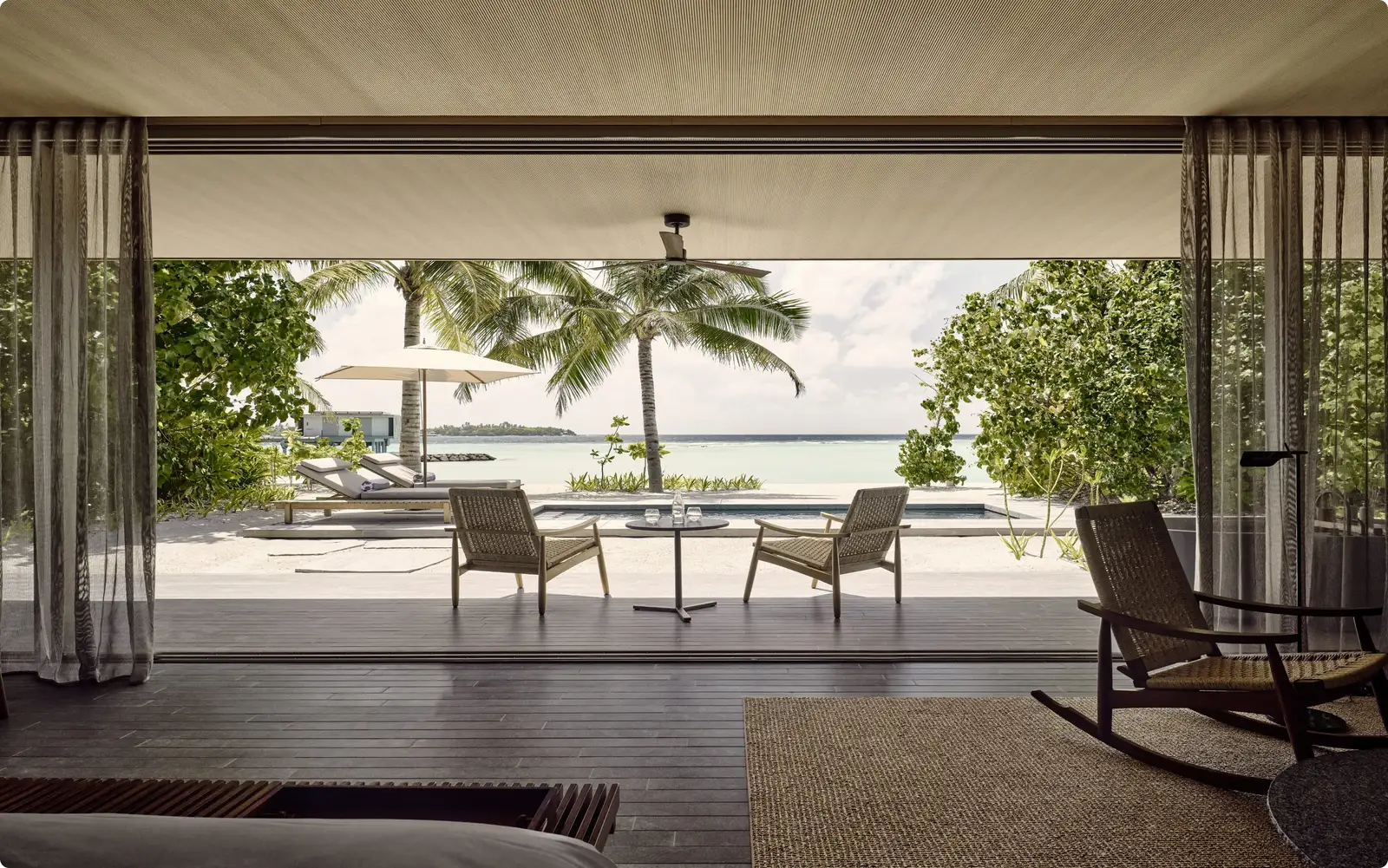 A beach villa with its glass open onto the deck, rocking chairs and the sea behind the palms