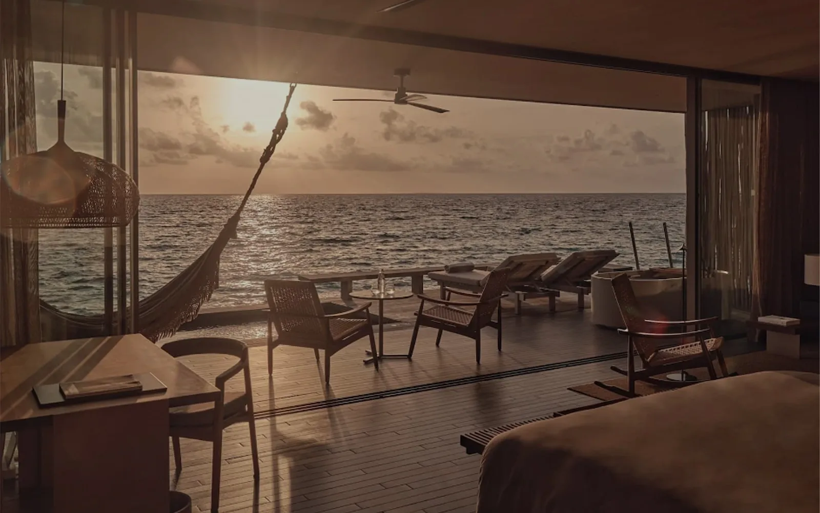 Sunset through the open glass of a water villa: the hammock, the chairs on the deck and the sea