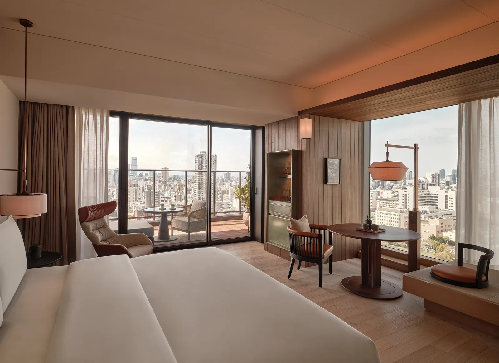 A Premier Room: a bed, armchairs and a table by a glass wall onto a balcony and the city