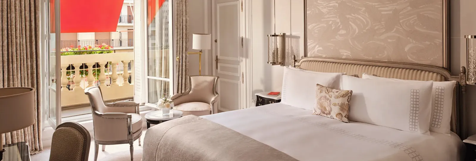 A bedroom with a bed, 2 pale armchairs and a table beside the French doors