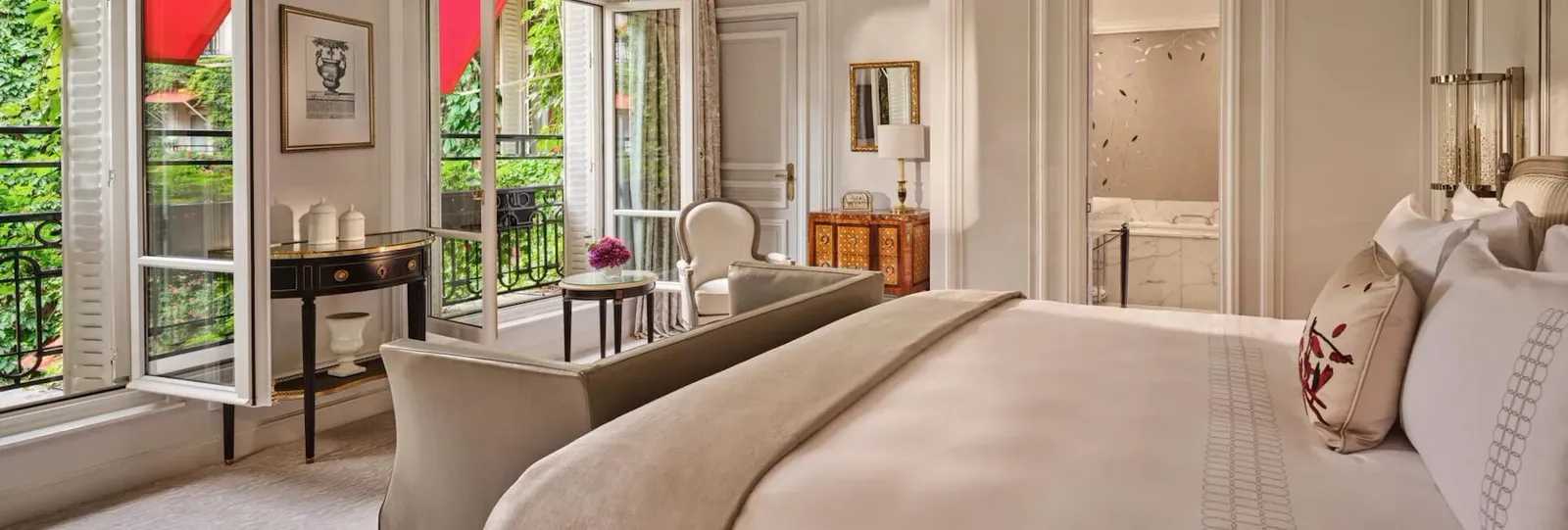A bedroom with a bed, pale armchair, lamps and open French doors