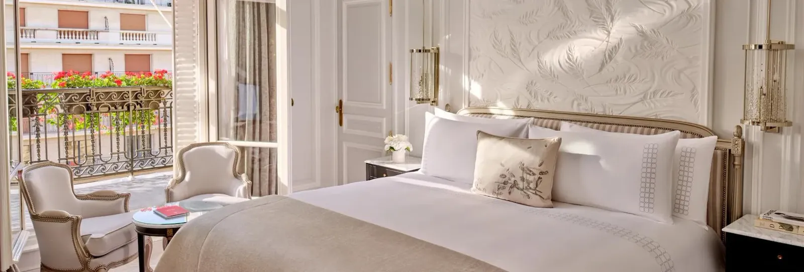 A bedroom with a bed, 2 pale armchairs and open French doors facing a flower-lined railing
