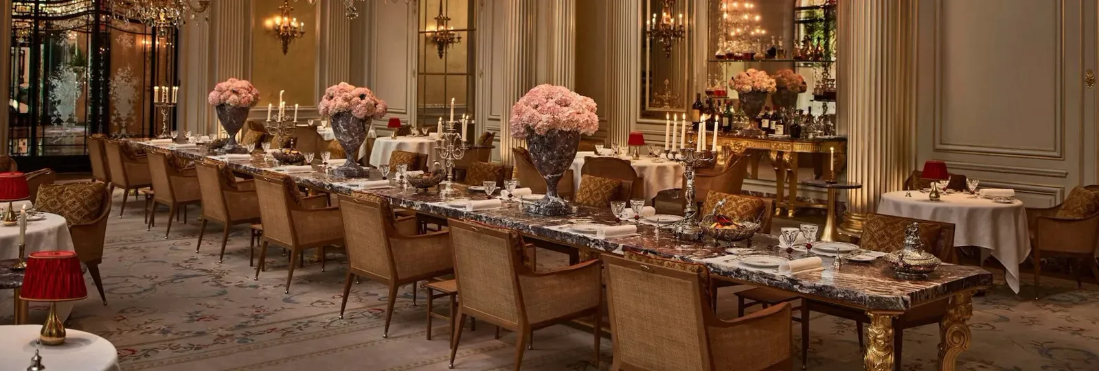 A dining room with a long laid table, pink flowers, candles and red lamps