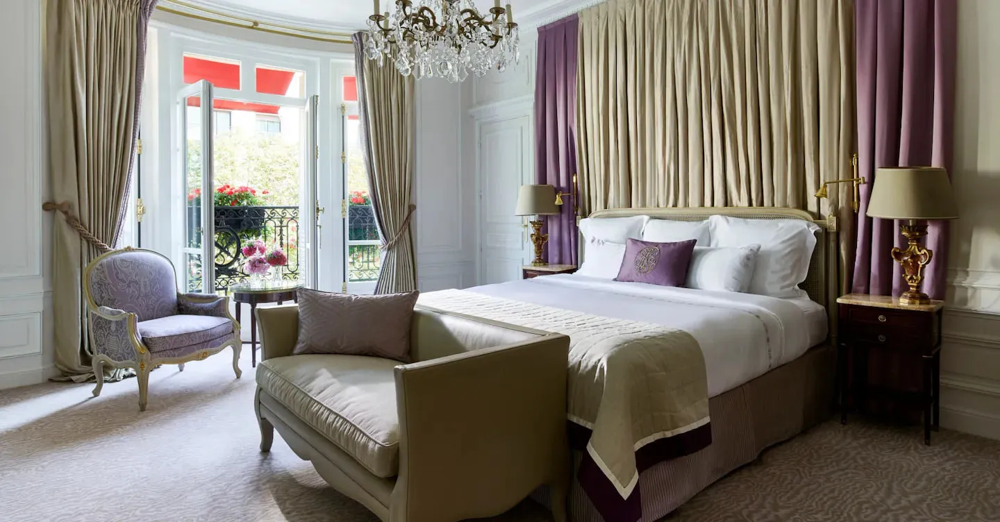 A bedroom with a bed, pale sofa, purple armchair and chandelier beside French doors