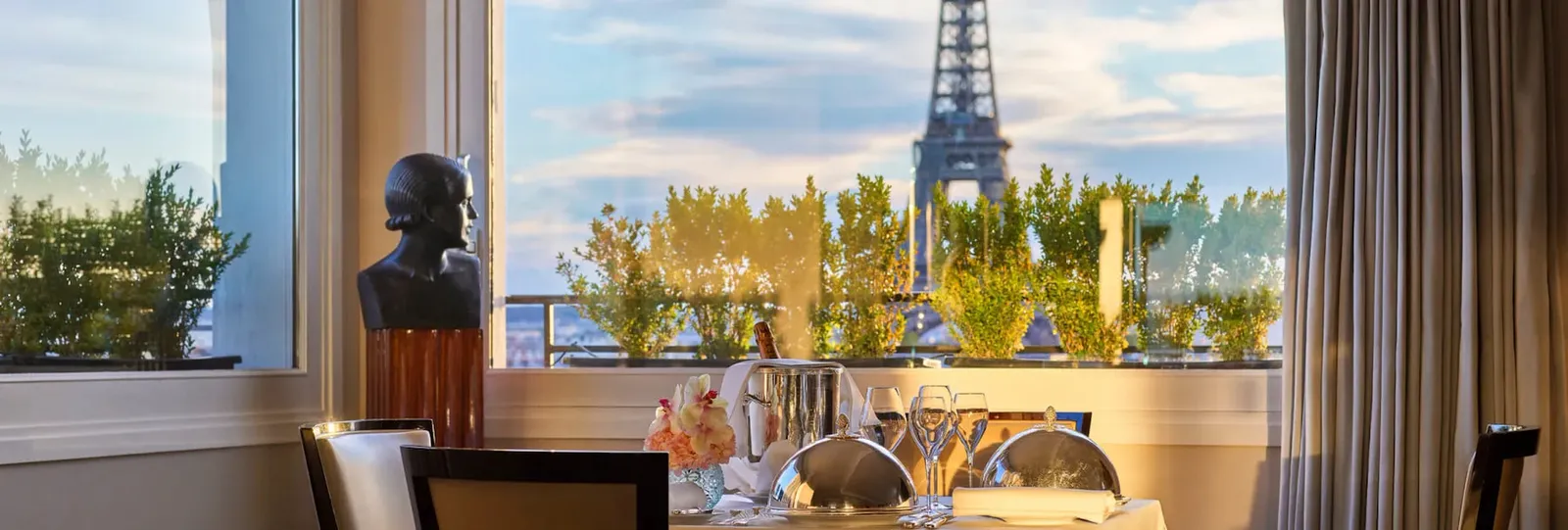 A table and 2 chairs by the window, with the Eiffel Tower beyond the trees