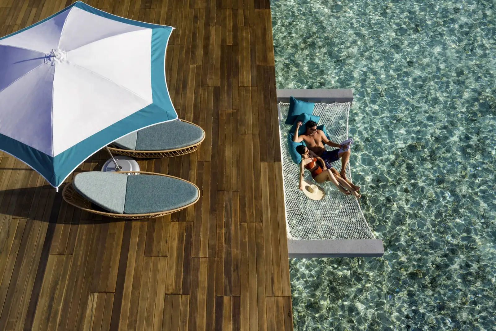 A couple in the hammock slung over the lagoon beside a water villa’s deck.