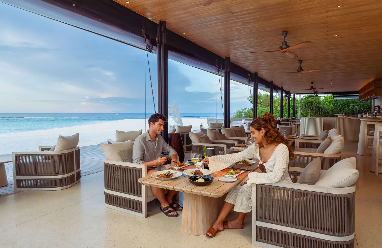 A couple at lunch on AMPERS&ND’s covered terrace, with woven armchairs and the beach beyond the glass.