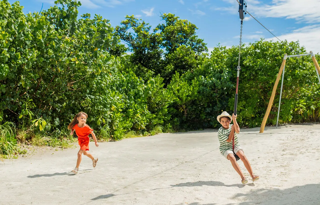 2 children playing on the sand at the Discovery Den, one riding the zip line among the trees.