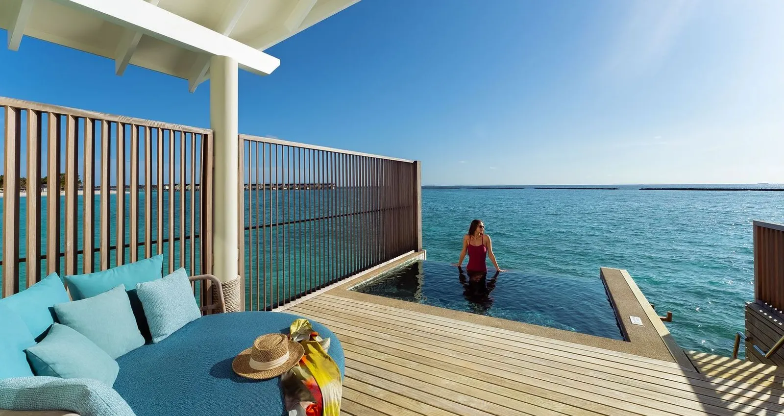 A Water Villa with Pool’s deck, with the blue sofa and a guest in the pool on the edge of the lagoon.