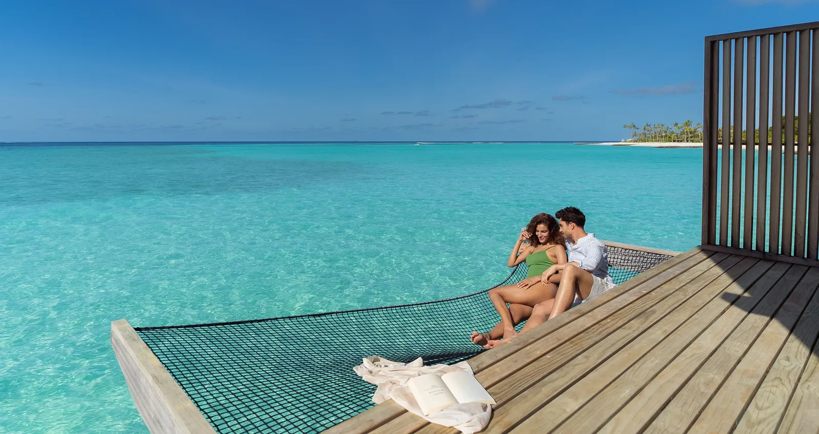 A couple lying on the net slung over the lagoon at the edge of a water villa’s deck.