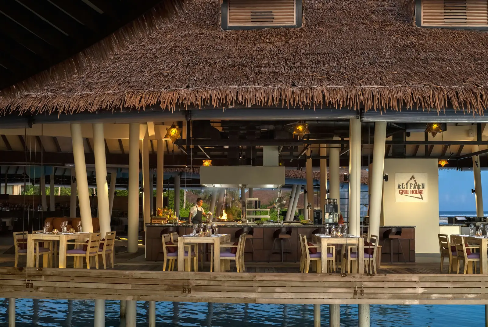 Alifaan over the water, under its thatch, with the open kitchen and laid tables.