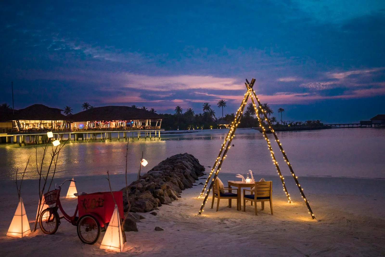 Dinner for 2 on the beach at dusk, under a frame of lights, with lanterns on the sand.