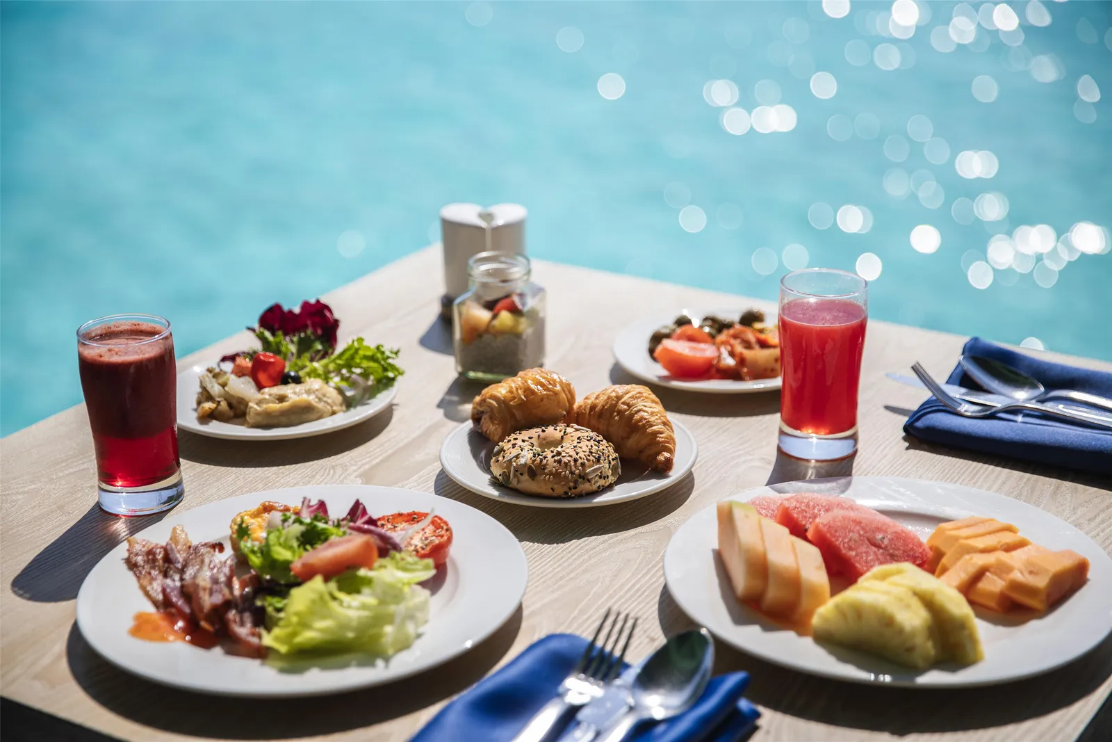 Breakfast on a table at Raha: croissants, fruit, bacon and juices, the sea glittering behind.