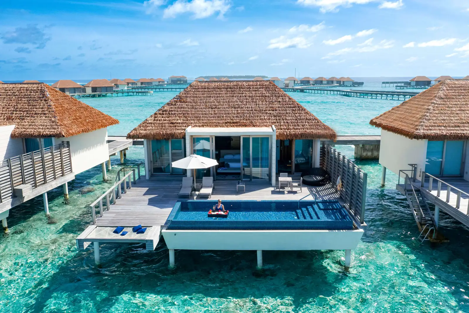 An overwater villa from above, with a floating breakfast in the pool on its deck.