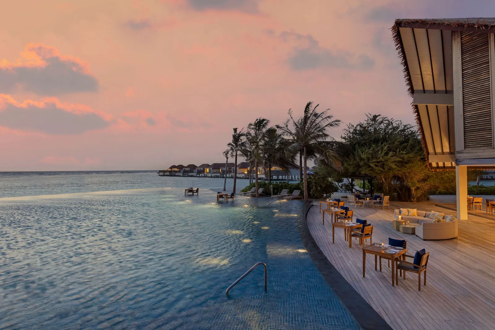 The Eats & Beats infinity pool at sunset, tables along its edge, palms and the overwater villas on the horizon.